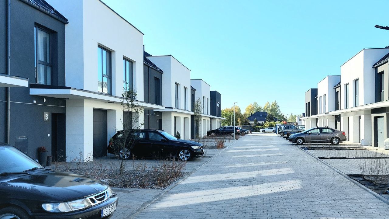 Nowoczesne szeregowe domy jednorodzinne z płaskimi dachami, pomalowane na biało i szaro, z samochodami zaparkowanymi wzdłuż wybrukowanej drogi dojazdowej.