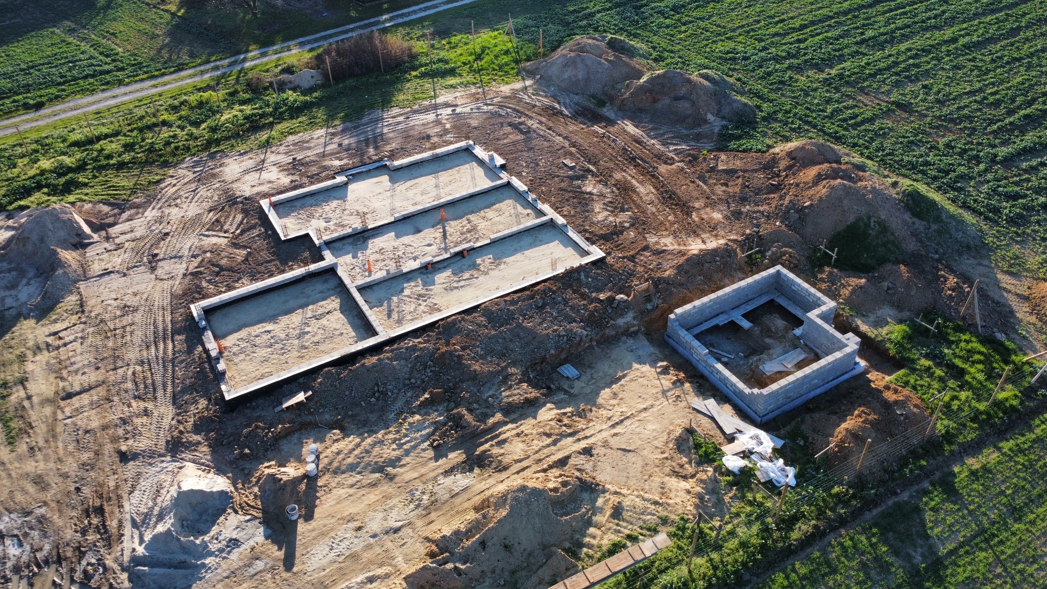 Widok z lotu ptaka na plac budowy z wykopanymi fundamentami domu, częściowo wylana płyta fundamentowa z widocznym zbrojeniem, obok mniejszy budynek z bloczków betonowych, teren otoczony polami...