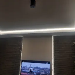 Nowoczesne oświetlenie sufitowe LED w narożniku pomieszczenia, ceglana ściana po bokach, telewizor z widokiem samolotu na dole.