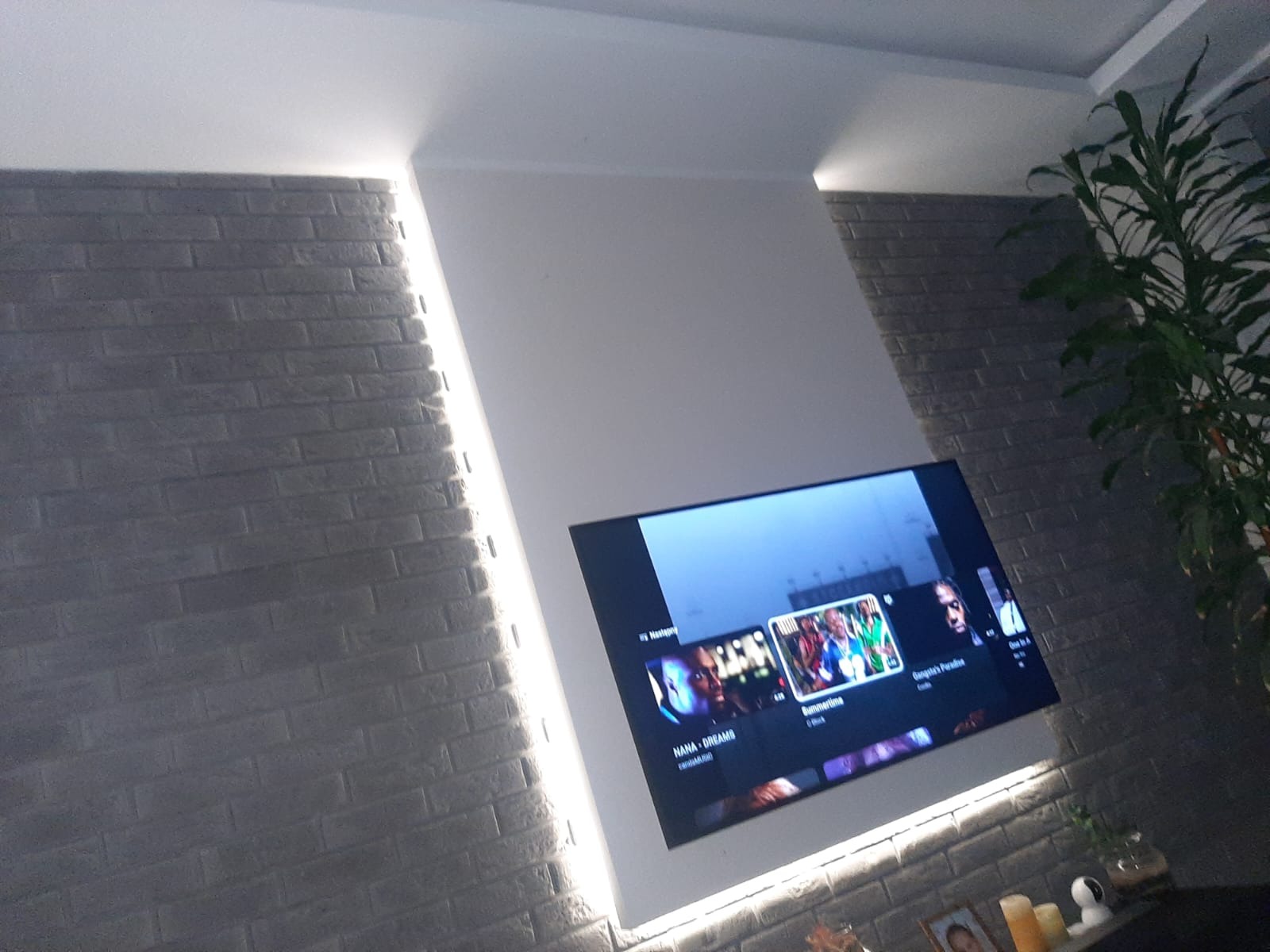 Telewizor zawieszony na ścianie z cegły, podświetlony listwami LED, obok roślina doniczkowa