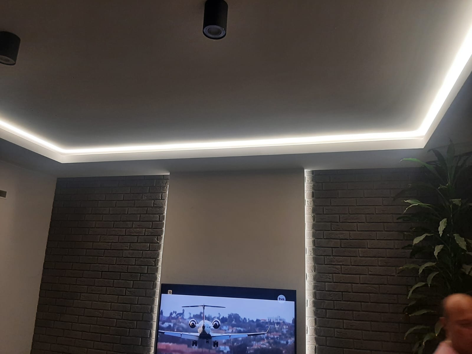 Nowoczesne oświetlenie sufitowe LED w narożniku pomieszczenia, ceglana ściana po bokach, telewizor z widokiem samolotu na dole.