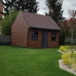 Niewielki drewniany budynek gospodarczy z brązowym dachem i szarymi oknami oraz drzwiami, otoczony zadbanym trawnikiem i ozdobnymi krzewami, w tle widać budynek z czerwoną dachówką i wieżą.