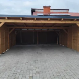 Drewniana wiata garażowa z płaskim dachem i ażurowymi ścianami bocznymi, widok od frontu na kostkę brukową.