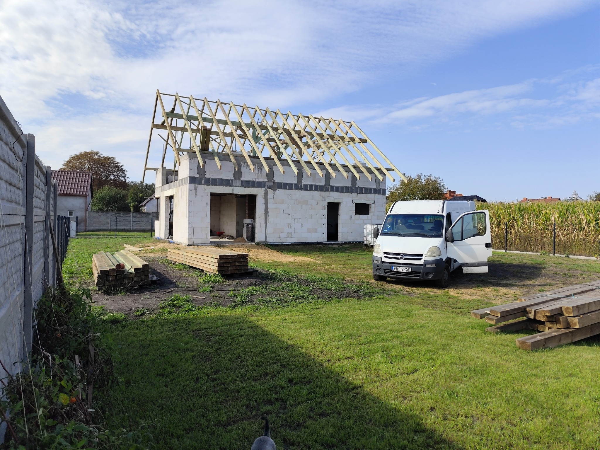 Budowa domu jednorodzinnego w stanie surowym otwartym z widoczną konstrukcją drewnianą dachu, obok zaparkowany biały bus dostawczy z otwartymi drzwiami.