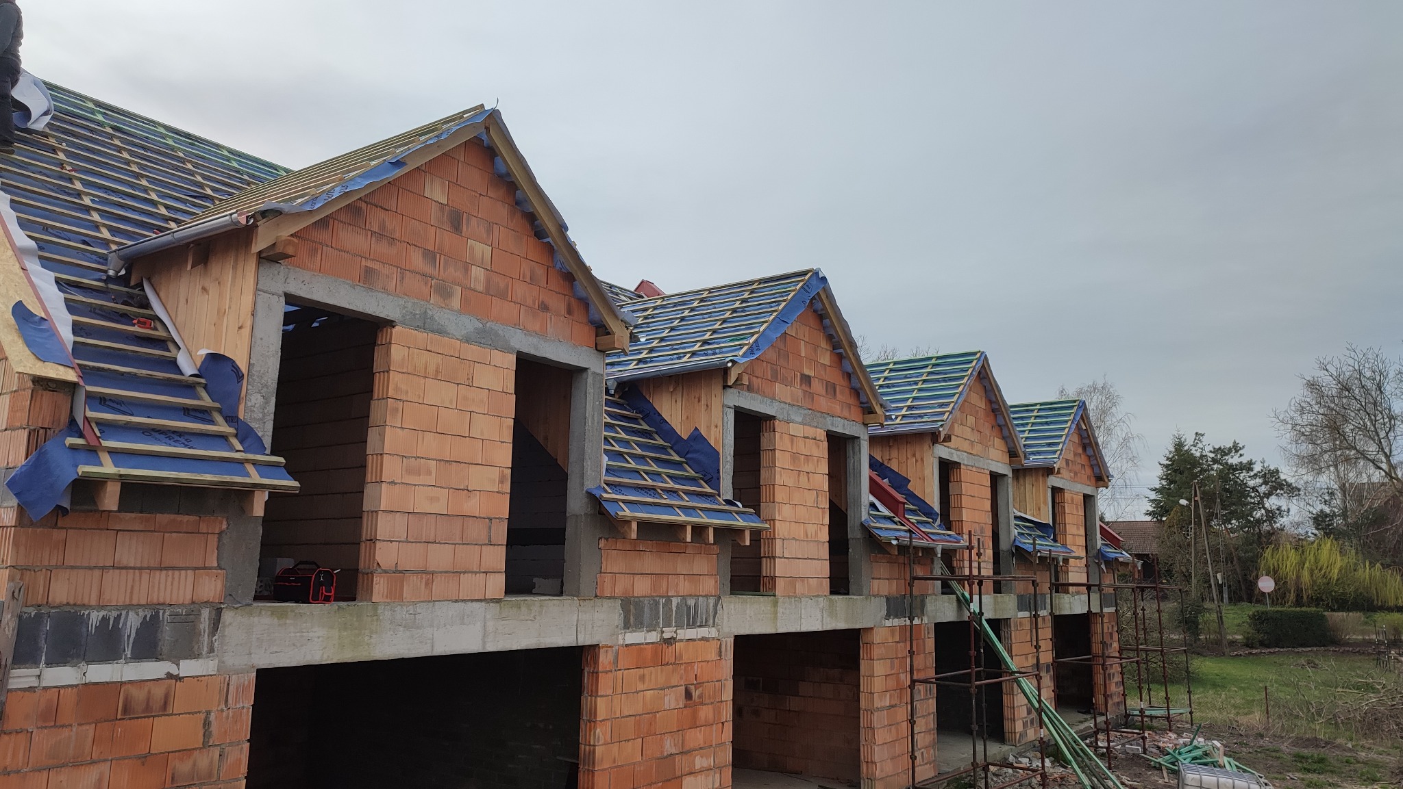 Budowa bliźniaczych domów z czerwonej cegły, widoczna więźba dachowa pokryta niebieską membraną, etap prac przed kryciem dachu, narzędzia na pierwszym planie.