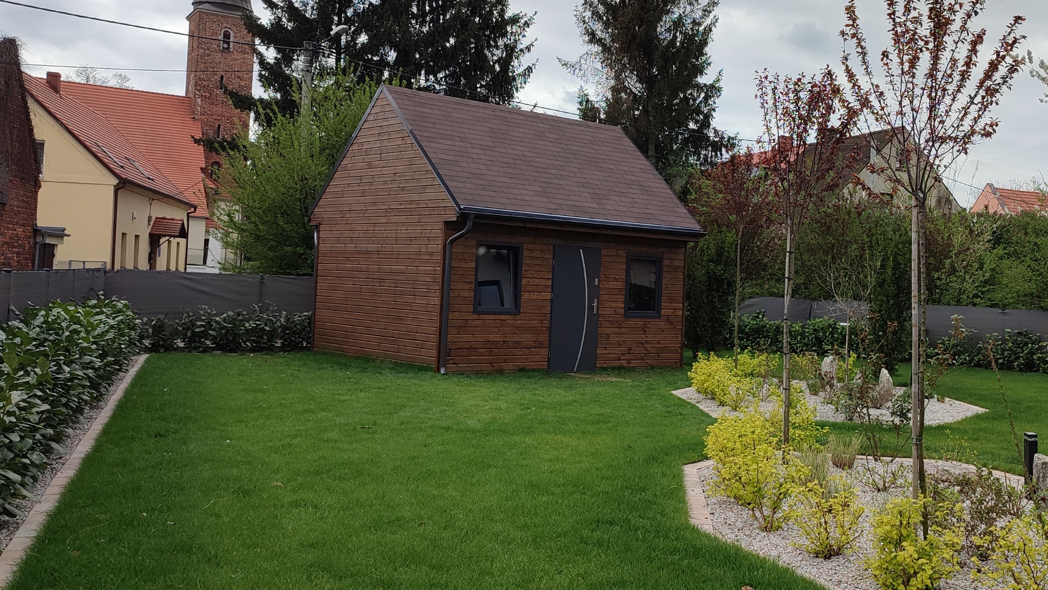 Niewielki drewniany budynek gospodarczy z brązowym dachem i szarymi oknami oraz drzwiami, otoczony zadbanym trawnikiem i ozdobnymi krzewami, w tle widać budynek z czerwoną dachówką i wieżą.