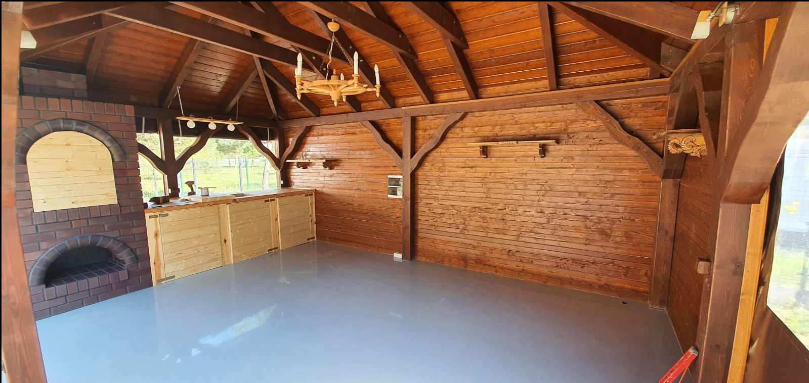 Wnętrze drewnianej altany z murowanym grillem i drewnianą zabudową kuchenną, oświetlone drewnianym żyrandolem.
