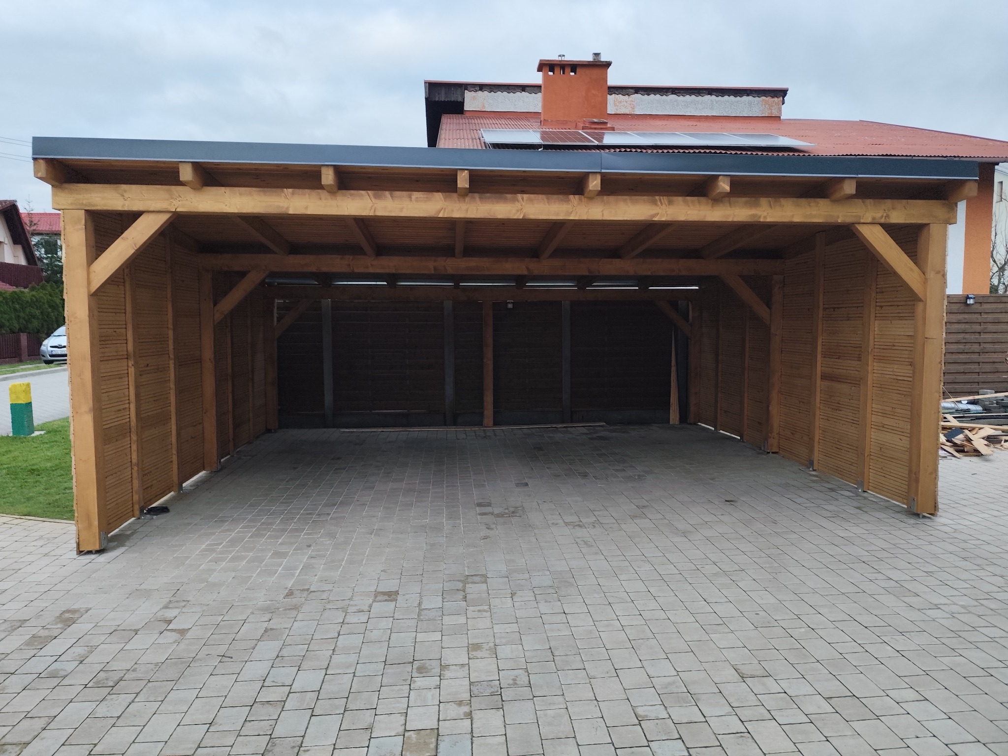 Drewniana wiata garażowa z płaskim dachem i ażurowymi ścianami bocznymi, widok od frontu na kostkę brukową.