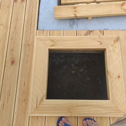 LuxBud Vladyslav Sirobaba - Drewniany taras z otworem rewizyjnym w ramie, widok z góry, fragment stopy w klapkach. Jasne drewno, równoległe deski, wkręty.