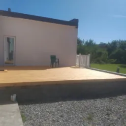 Nowy drewniany taras przy domu z jasną elewacją i zielonym krzesłem. Świeżo ułożone deski tarasowe, widoczne detale wykończenia. Słoneczny dzień.