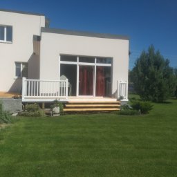 LuxBud Vladyslav Sirobaba - Nowoczesny dom z tarasem i schodami z jasnego drewna, otoczony zielonym trawnikiem i roślinnością. Widok z przodu, słoneczny dzień.