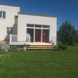 Nowoczesny dom z tarasem i schodami z jasnego drewna, otoczony zielonym trawnikiem i roślinnością. Widok z przodu, słoneczny dzień.