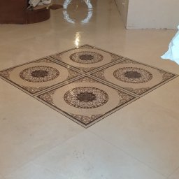 LuxBud Vladyslav Sirobaba - Połyskująca podłoga wyłożona jasnymi płytkami z dekoracyjnym motywem w kształcie rombu i okręgów. Widoczny fragment schodów i drzwi wejściowe.