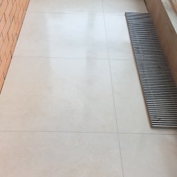 LuxBud Vladyslav Sirobaba - Jasne, kremowe płytki podłogowe ułożone na balkonie, widoczna ceglana ściana, okno i narzędzia glazurnicze, kratka ściekowa. Ukończone prace glazurnicze.