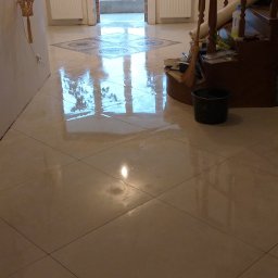 LuxBud Vladyslav Sirobaba - Połyskująca, kremowa podłoga z płytek ułożonych w karo, z dekoracyjnym wzorem przy oknie. Widoczne schody, wiadro i akcesoria. Świeże wykończenie wnętrza.