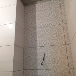 LuxBud Vladyslav Sirobaba - Fragment wykończonej ściany w łazience z mozaiką w odcieniach szarości i białymi płytkami na narożniku. Widoczny kabel wystający ze ściany i surowy betonowy sufit.