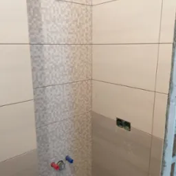 Wykończona ściana w łazience z beżowych płytek, z fragmentem dekoracyjnej mozaiki i widocznymi przyłączami wodnymi. W tle surowy beton i instalacja elektryczna.