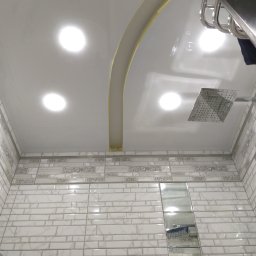 LuxBud Vladyslav Sirobaba - Biały sufit napinany z oświetleniem i dekoracyjnym łukiem w łazience, wykończonej jasnymi płytkami z dekoracyjnym pasem. Widoczna deszczownica.