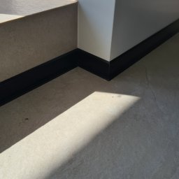 LuxBud Vladyslav Sirobaba - Światło słoneczne na beżowej podłodze z płytek, czarne listwy przypodłogowe przy białej ścianie i schodach. Minimalizm, geometria.