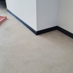 LuxBud Vladyslav Sirobaba - Ściana z czarnymi listwami przypodłogowymi i beżowymi płytkami podłogowymi. Widoczny narożnik i fragment różowego ręcznika.