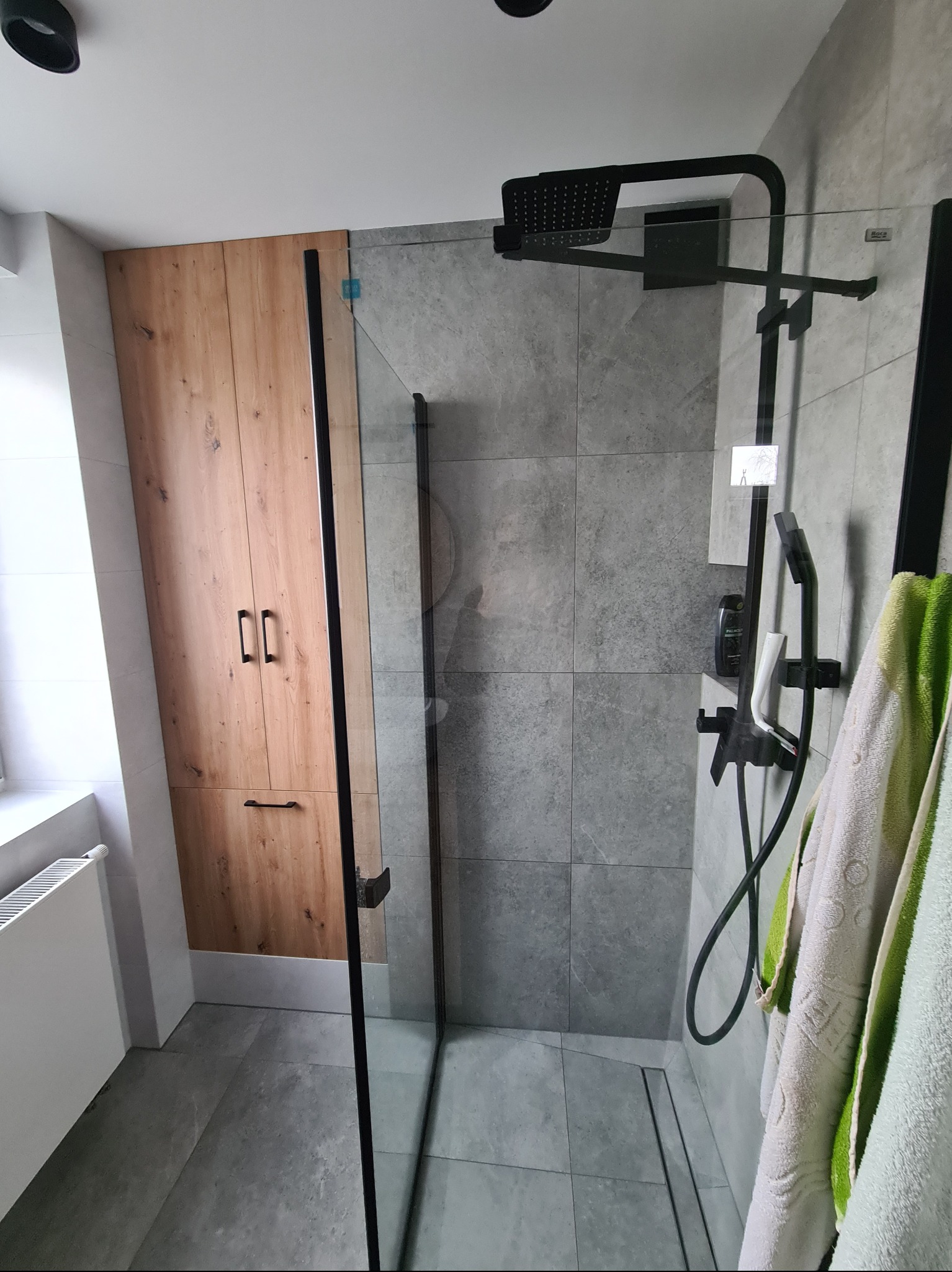 Nowoczesna łazienka z kabiną prysznicową typu walk-in, szare płytki, drewniana szafka i czarna armatura. Minimalistyczny design z akcentem naturalnego drewna.