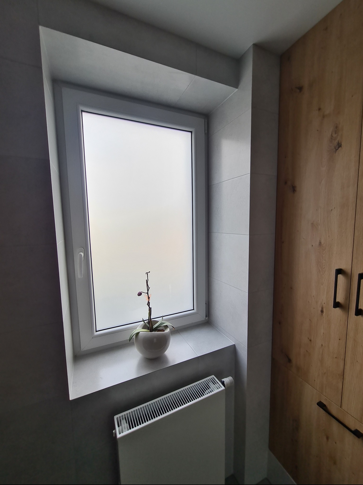 Wykończona łazienka z oknem, na parapecie doniczka z kwiatem, obok drewniana szafka z czarnymi uchwytami. Szare płytki na ścianach i biały grzejnik. Nowoczesny design.