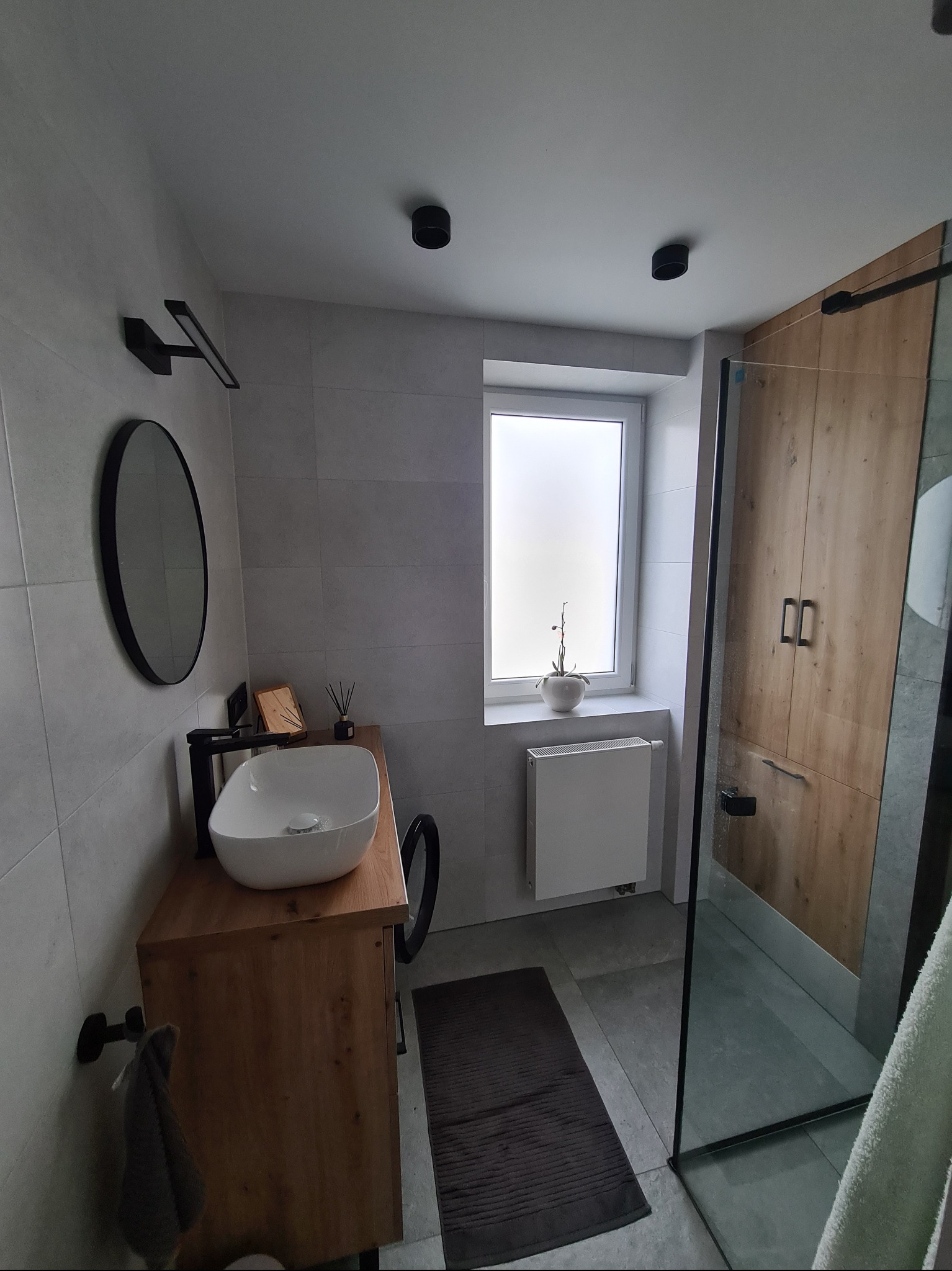 Nowoczesna łazienka z drewnianymi akcentami: szafka pod umywalką, kabina prysznicowa z drewnianymi drzwiami, okrągłe lustro i szare płytki.