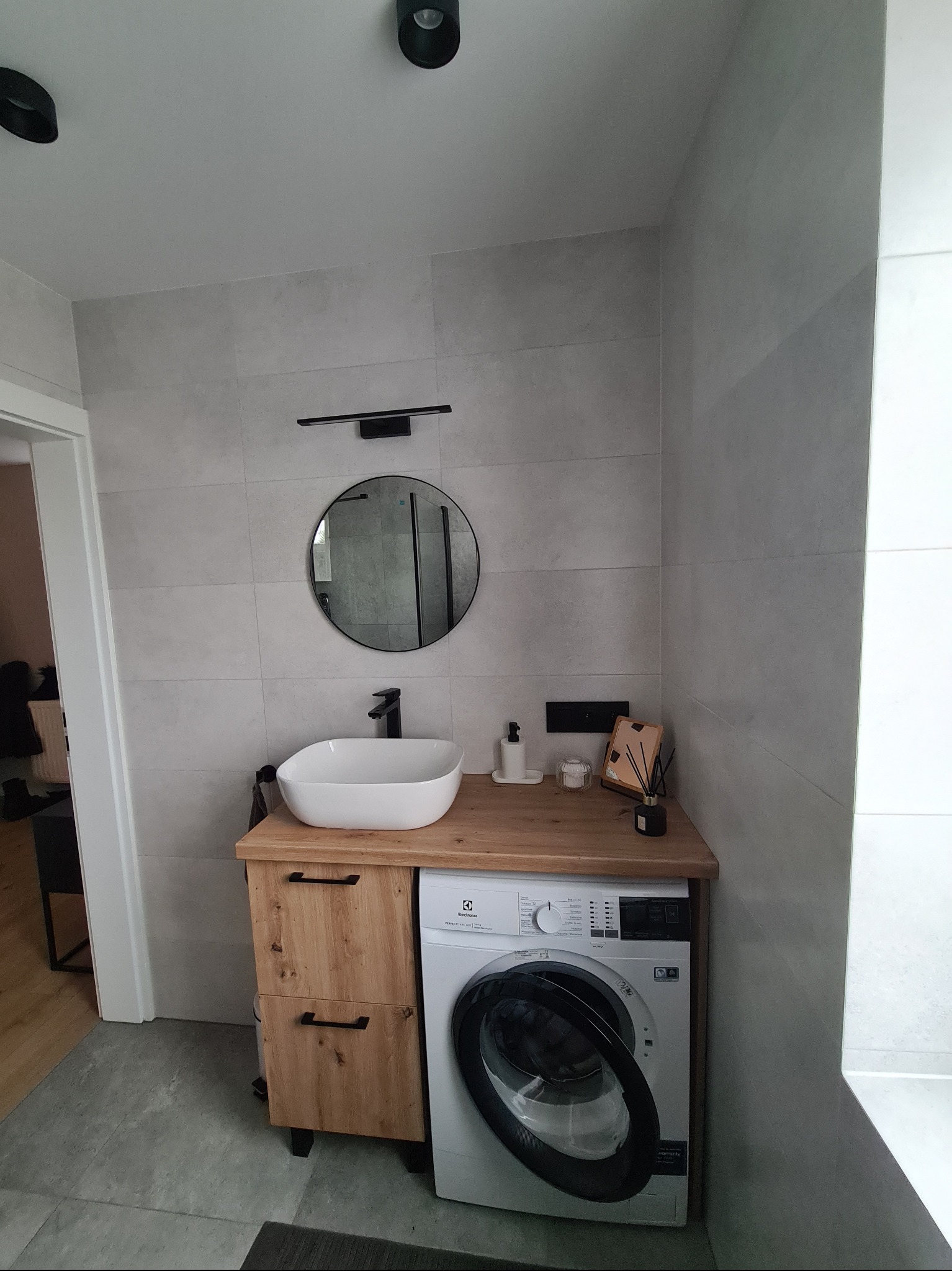 Nowoczesna łazienka z umywalką nablatową, okrągłym lustrem i pralką pod blatem. Minimalistyczny design, szare płytki, drewniane szafki i czarne akcenty oświetleniowe.