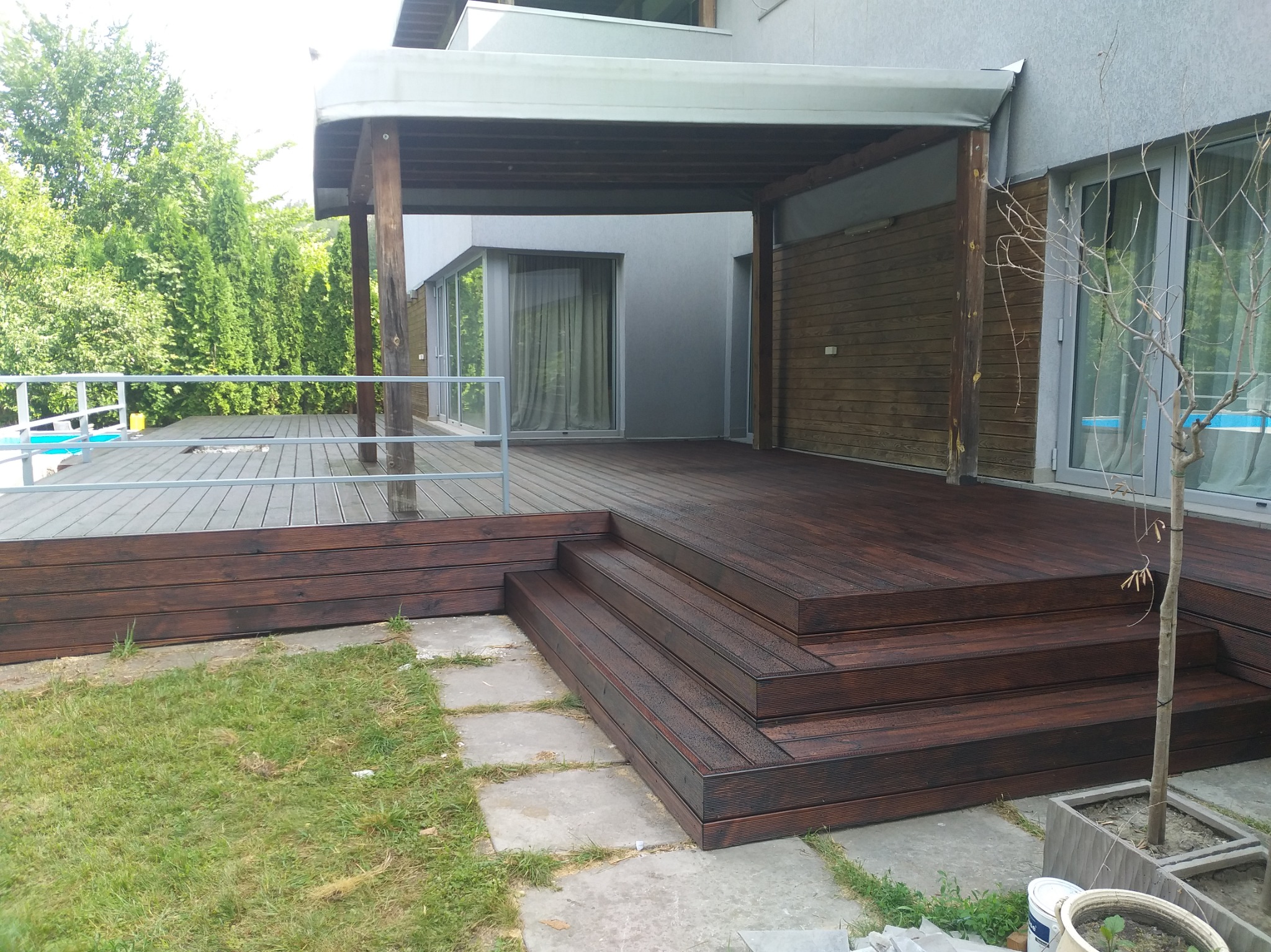 Ciemnobrązowy taras z drewna kompozytowego, schody z desek, zadaszenie, widok na zieleń i basen. Nowoczesne wykończenie zewnętrzne domu.
