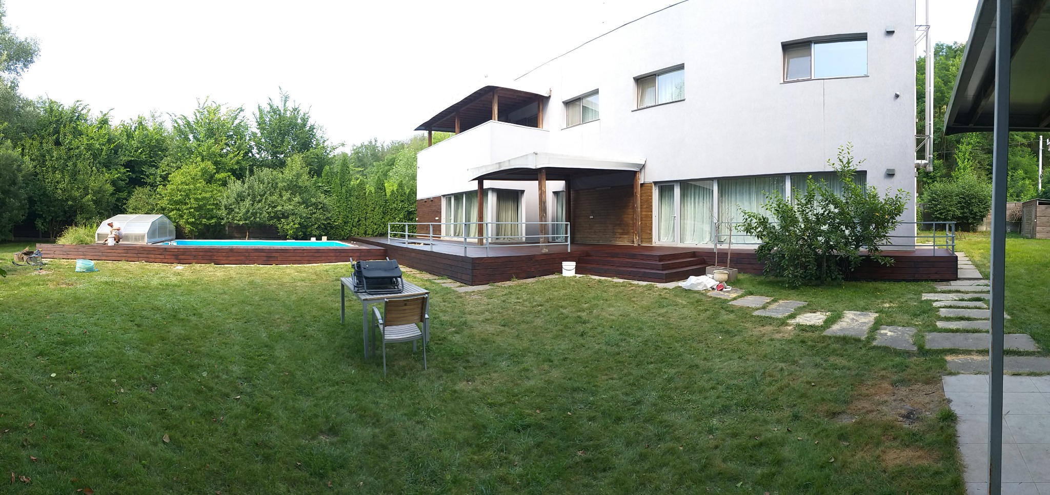 Nowoczesny dom z tarasem z drewna egzotycznego i basenem w ogrodzie. Widoczny fragment elewacji i otoczenie. Stolik z grillem na trawniku.