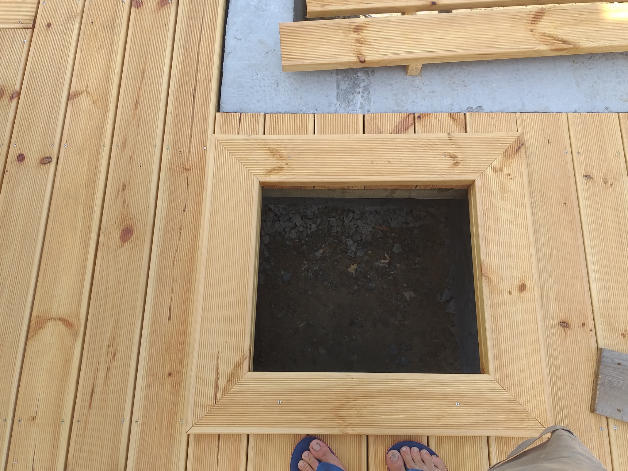 Drewniany taras z otworem rewizyjnym w ramie, widok z góry, fragment stopy w klapkach. Jasne drewno, równoległe deski, wkręty.