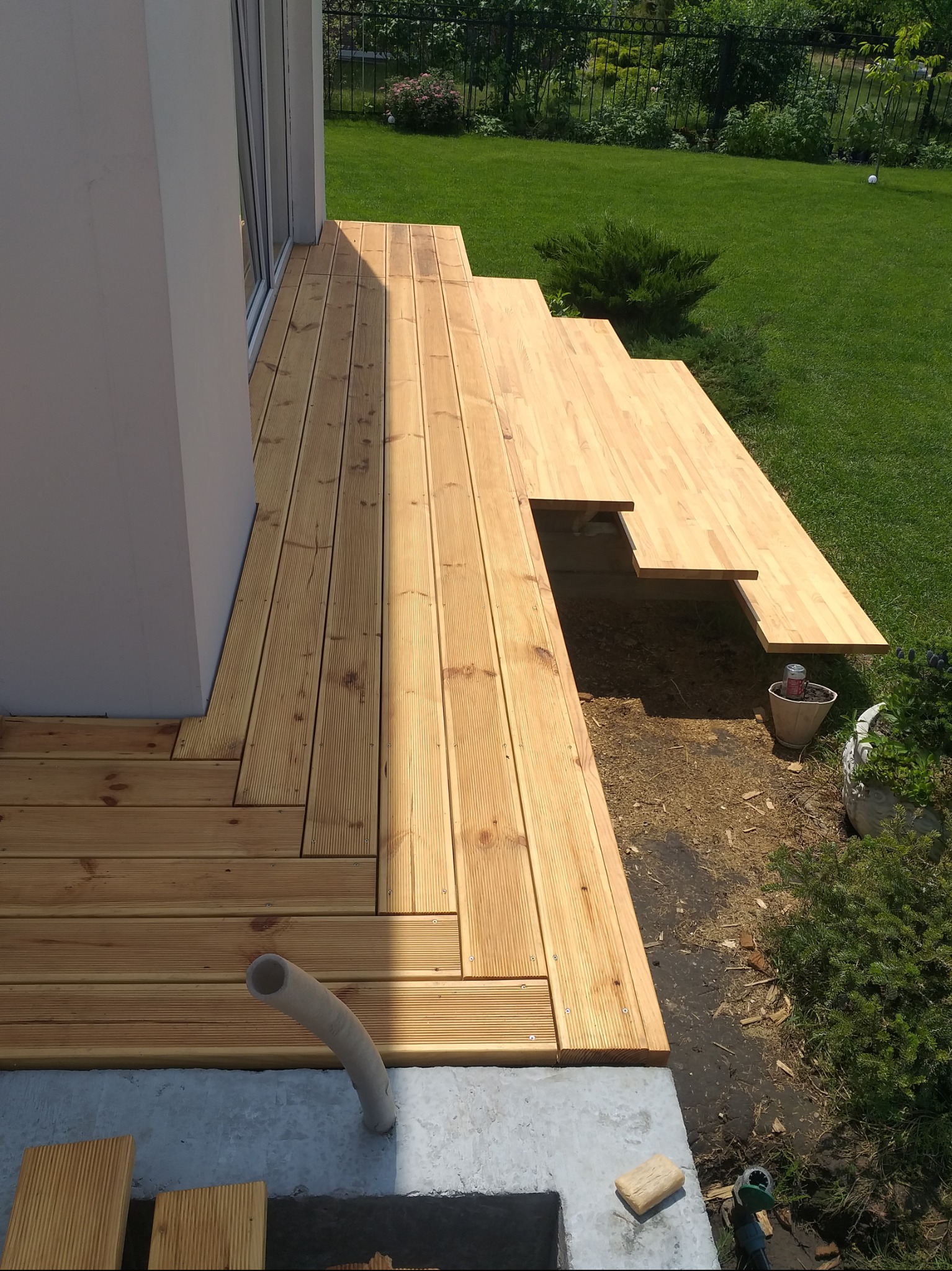 Drewniany taras w trakcie budowy, deski ułożone przy domu, widoczne elementy konstrukcyjne i narzędzia. Trawa i zieleń w tle.