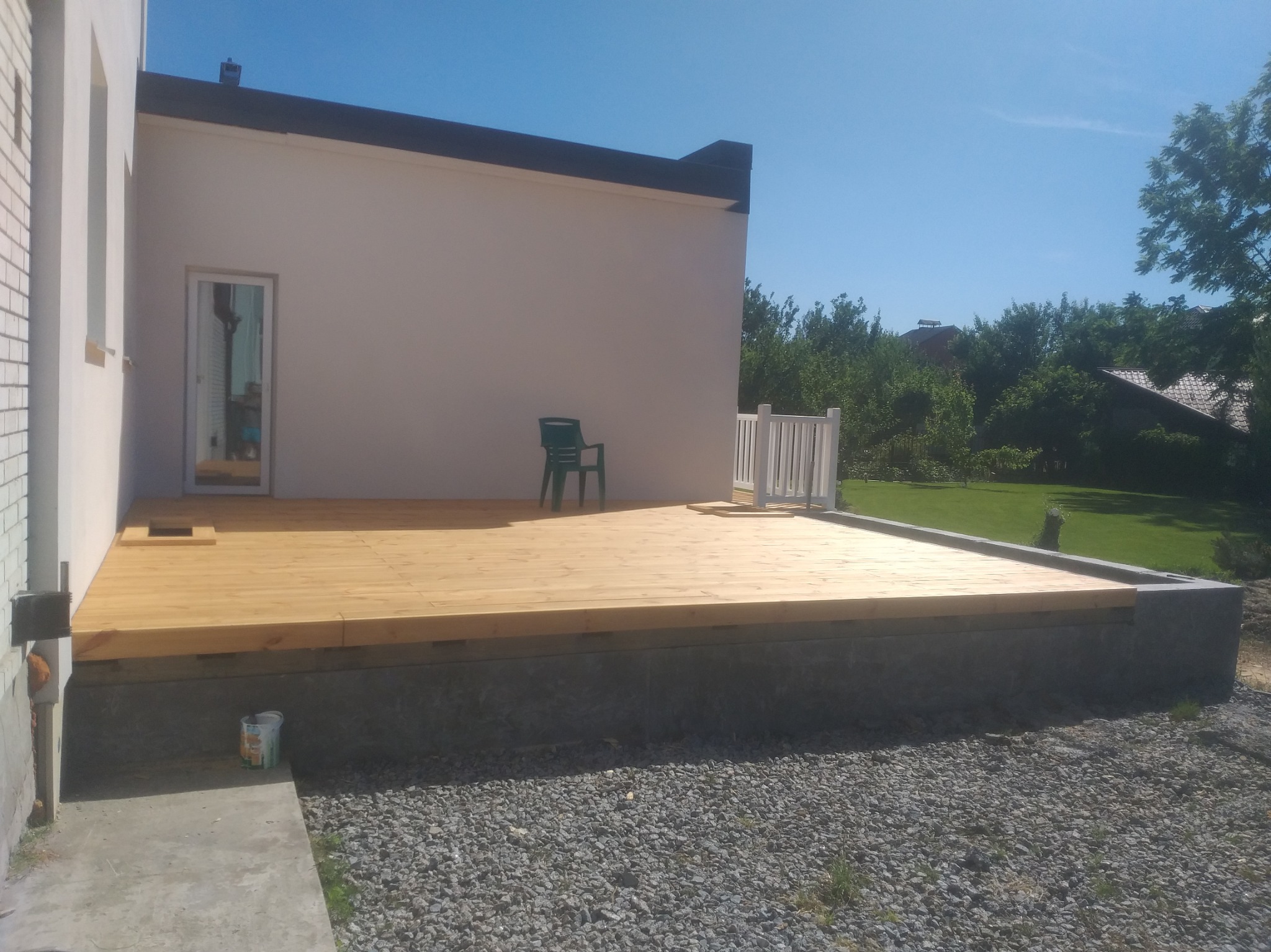 Nowy drewniany taras przy domu z jasną elewacją i zielonym krzesłem. Świeżo ułożone deski tarasowe, widoczne detale wykończenia. Słoneczny dzień.