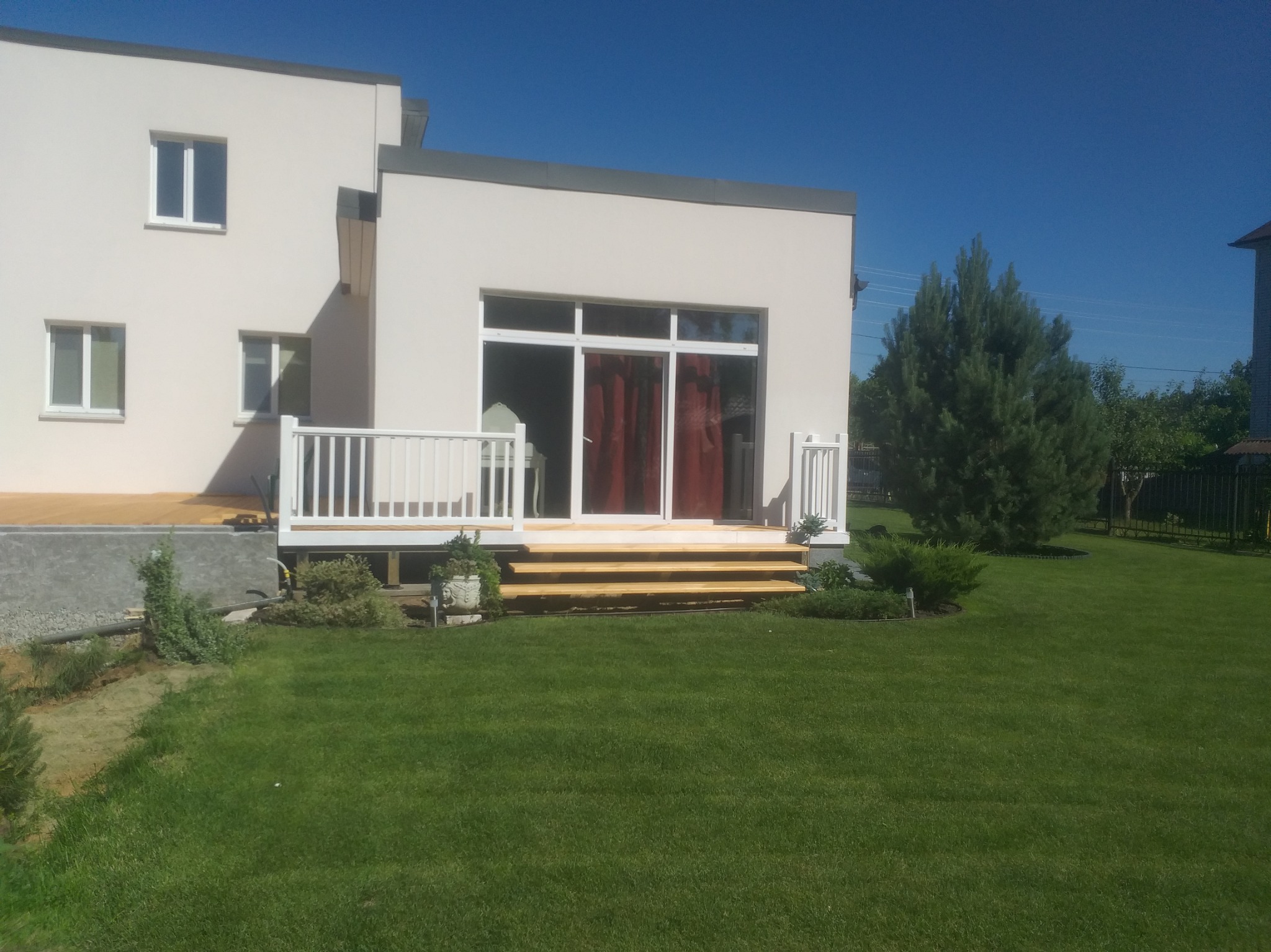 Nowoczesny dom z tarasem i schodami z jasnego drewna, otoczony zielonym trawnikiem i roślinnością. Widok z przodu, słoneczny dzień.