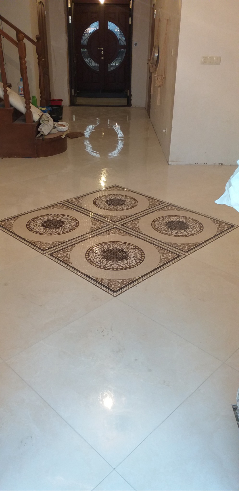 Połyskująca podłoga wyłożona jasnymi płytkami z dekoracyjnym motywem w kształcie rombu i okręgów. Widoczny fragment schodów i drzwi wejściowe.