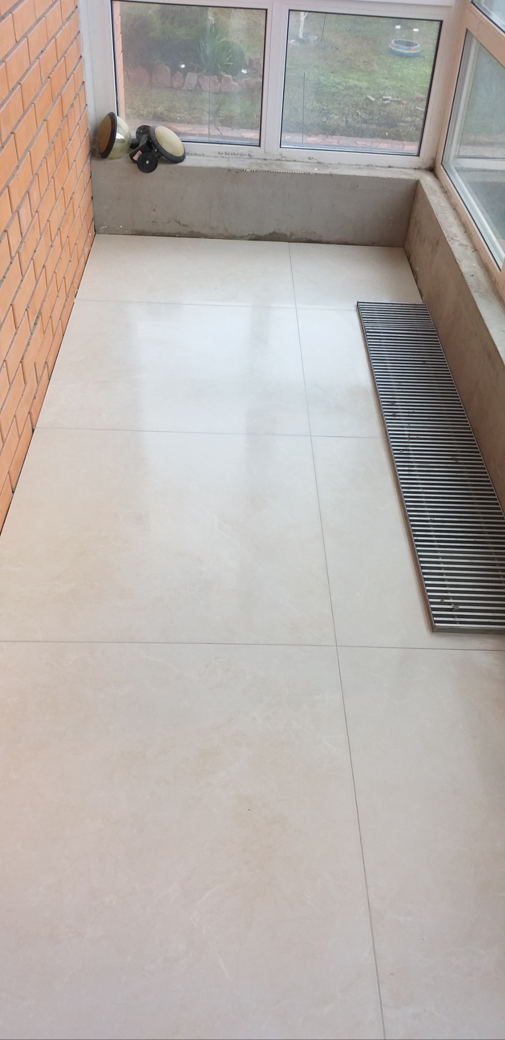 Jasne, kremowe płytki podłogowe ułożone na balkonie, widoczna ceglana ściana, okno i narzędzia glazurnicze, kratka ściekowa. Ukończone prace glazurnicze.