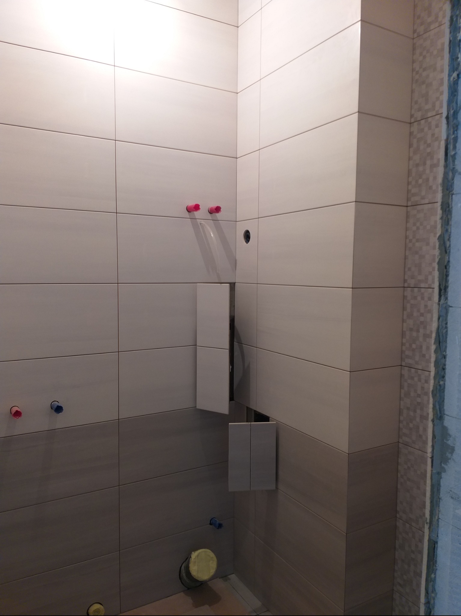 Ściana łazienki w trakcie wykańczania z beżowymi, poziomymi płytkami, widoczne instalacje wodne i fragment stelaża podtynkowego.