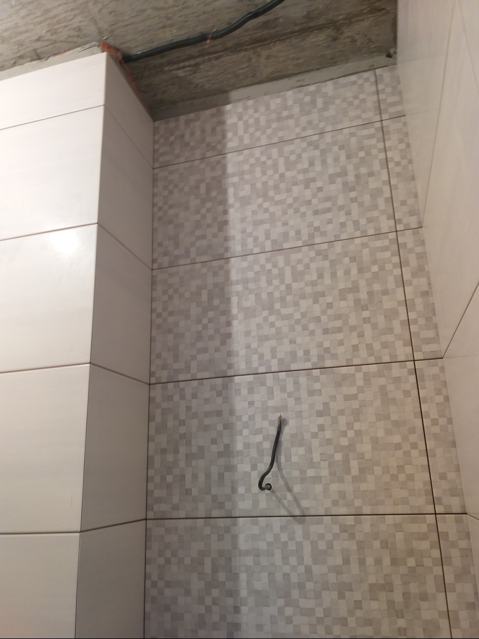 Fragment wykończonej ściany w łazience z mozaiką w odcieniach szarości i białymi płytkami na narożniku. Widoczny kabel wystający ze ściany i surowy betonowy sufit.