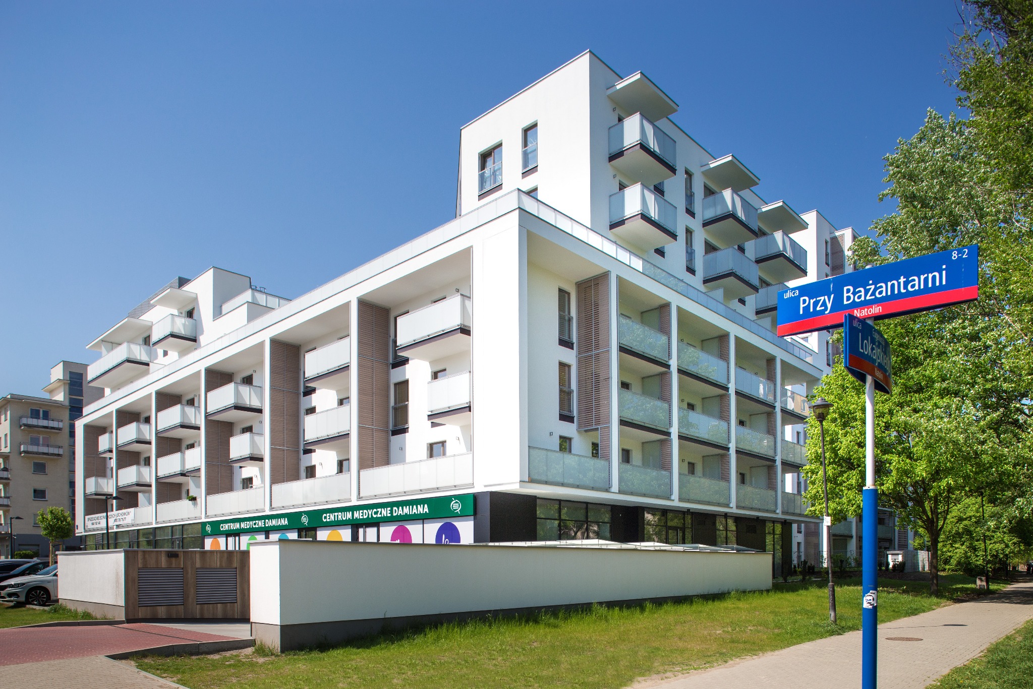 Nowoczesny, biały budynek apartamentowy z balkonami i szklanymi balustradami, widoczny znak uliczny 'Przy Bażantarni' i logo Centrum Medycznego Damiana na parterze, błękitne niebo w tle.