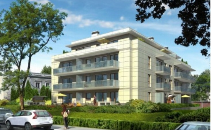 Nowoczesny, trzypiętrowy apartamentowiec z balkonami i szklanymi balustradami, otoczony zielenią i zaparkowanymi samochodami w słoneczny dzień. Widok z poziomu ulicy.