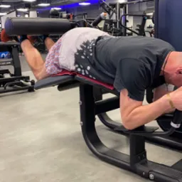 Mężczyzna ćwiczy mięśnie dwugłowe uda na maszynie w siłowni, ubrany w koszulkę z efektem tie-dye i wzorzyste spodenki, z widoczną butelką wody w tle.