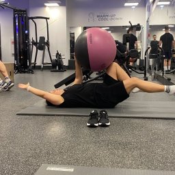 Adrian - Trener Personalny - Osoba ćwiczy z piłką fitness na macie w siłowni. Widoczne buty obok maty, lustra i sprzęt do ćwiczeń w tle. Napis 'Strefa Warm-up Cool-down' na ścianie.