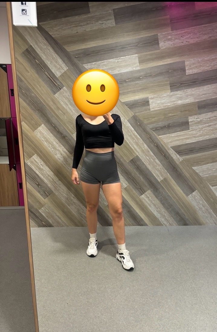 Osoba w stroju sportowym (czarne legginsy, top z długim rękawem, białe skarpetki i buty) stoi na szarym tle, z drewnianą ścianą w tle.