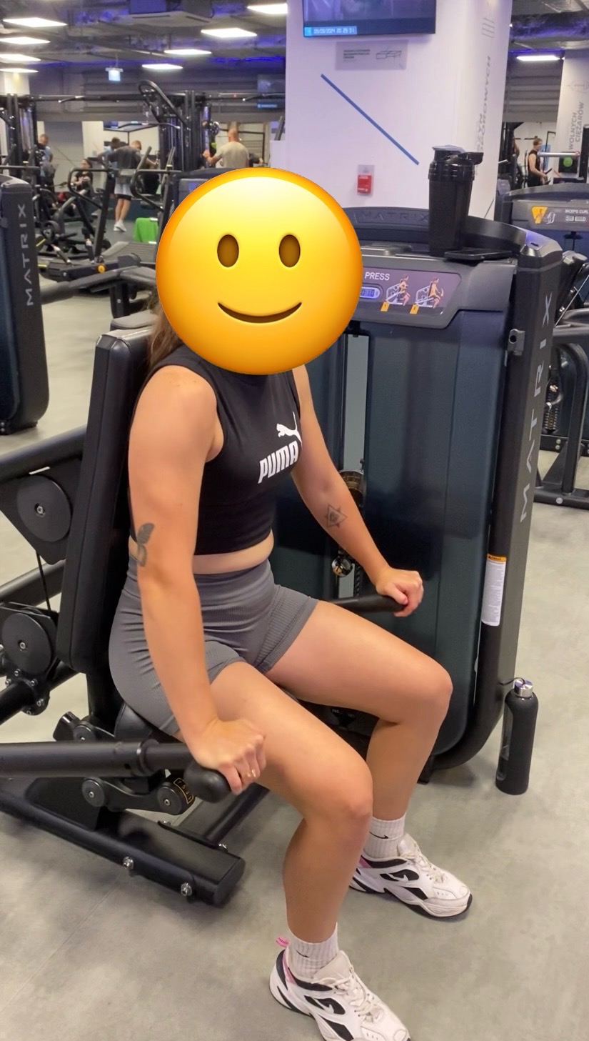 Kobieta ćwiczy na maszynie do wyciskania nogami w nowoczesnej siłowni, widoczny tatuaż na ramieniu i butelka z wodą obok maszyny.