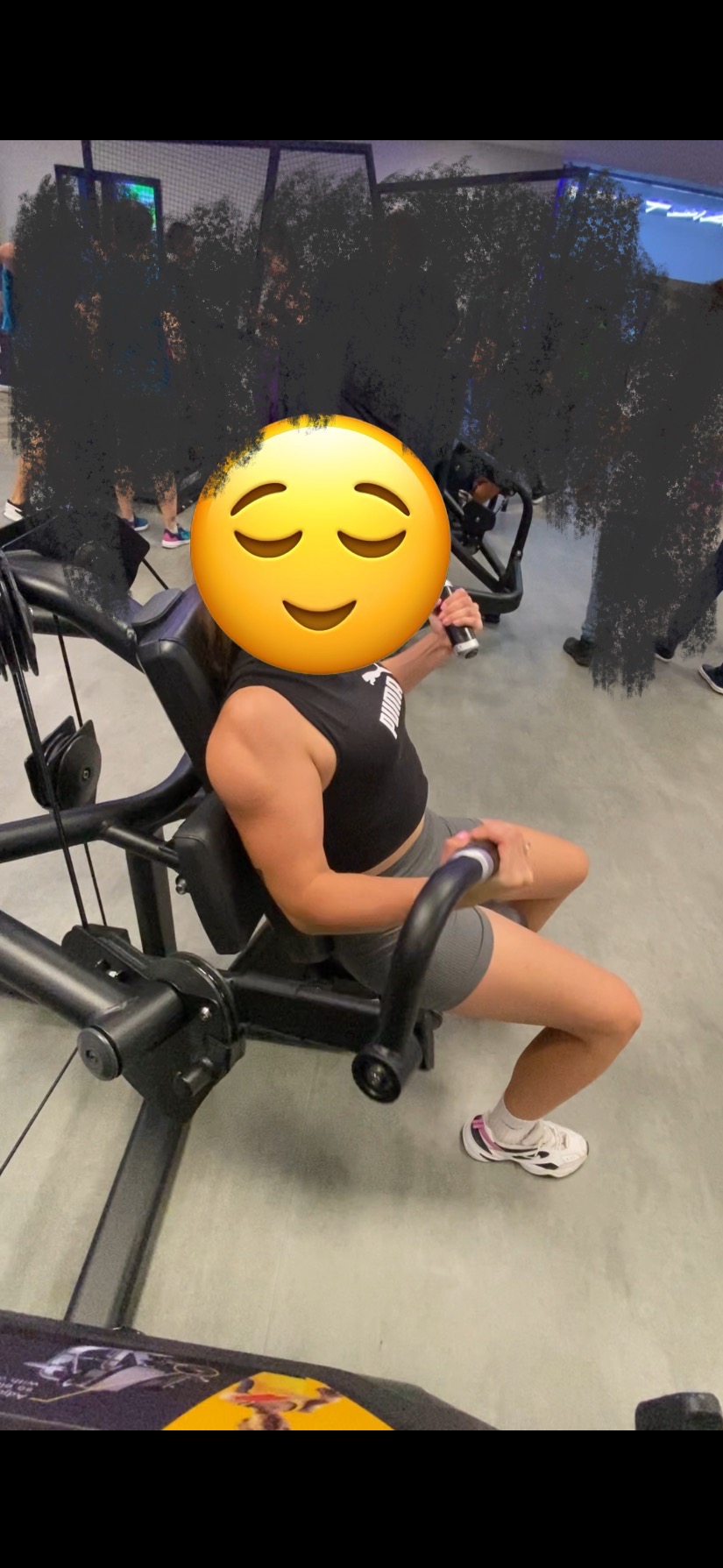 Kobieta ćwiczy na maszynie do wyciskania na klatkę piersiową w siłowni, widoczne mocno zarysowane mięśnie ramion, na podłodze naklejka z grafiką.