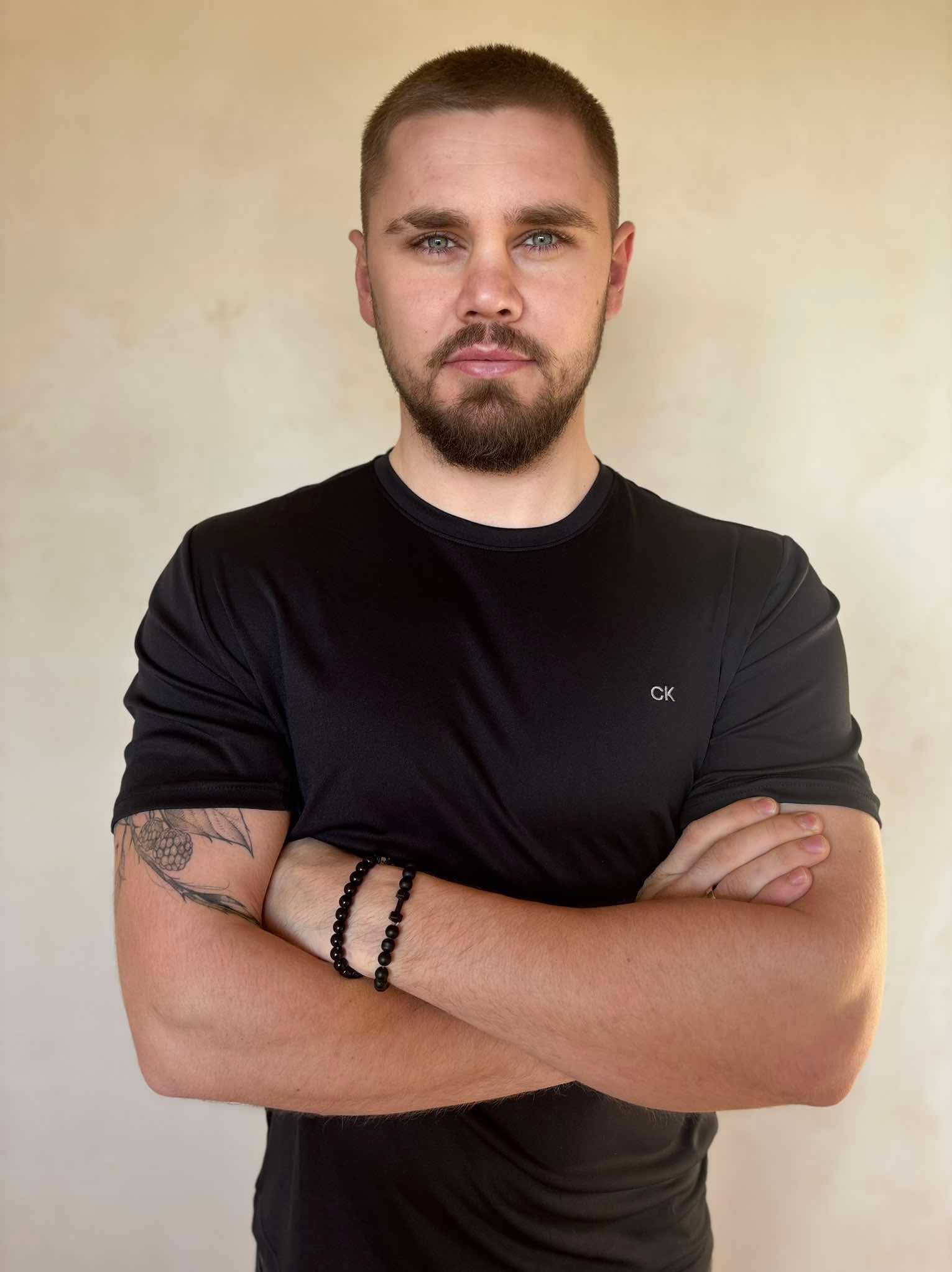 Portret mężczyzny z brodą i niebieskimi oczami, w czarnej koszulce CK, z tatuażem na ramieniu i czarną bransoletką z kamieni, na neutralnym tle.