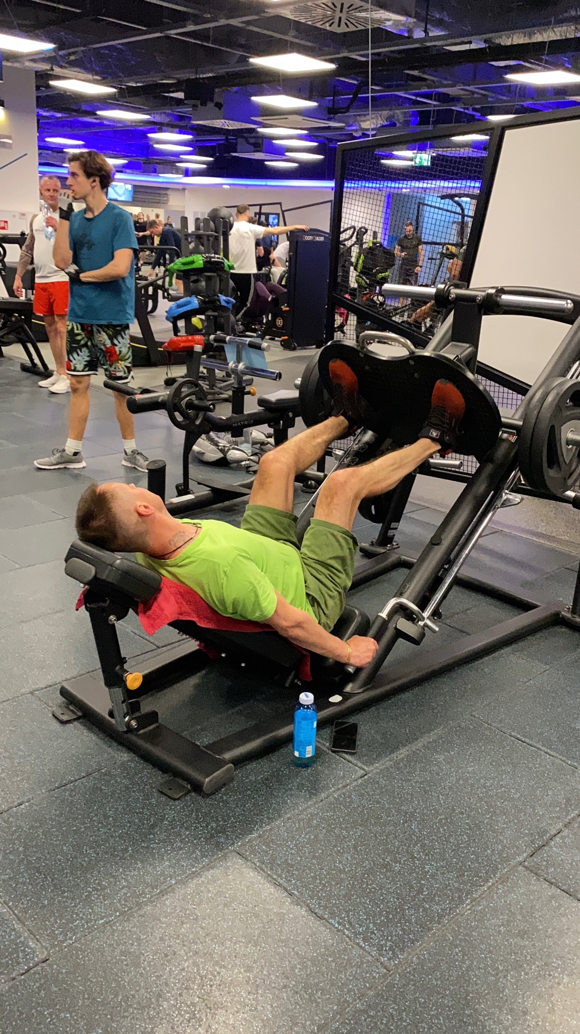 Mężczyzna w zielonej koszulce ćwiczy na suwnicy do nóg w siłowni w Gliwicach, obok butelka wody i telefon.