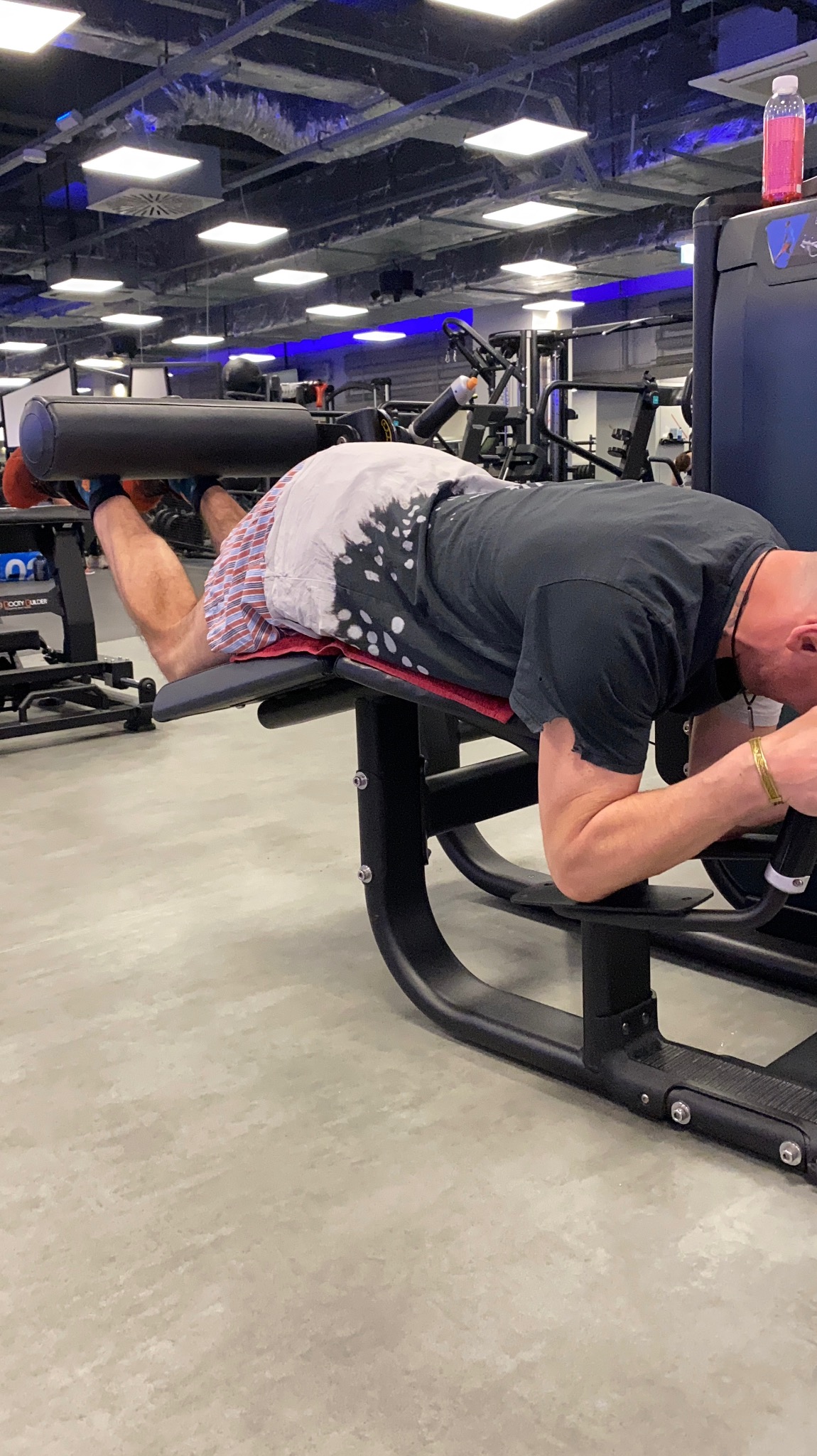 Mężczyzna ćwiczy mięśnie dwugłowe uda na maszynie w siłowni, ubrany w koszulkę z efektem tie-dye i wzorzyste spodenki, z widoczną butelką wody w tle.