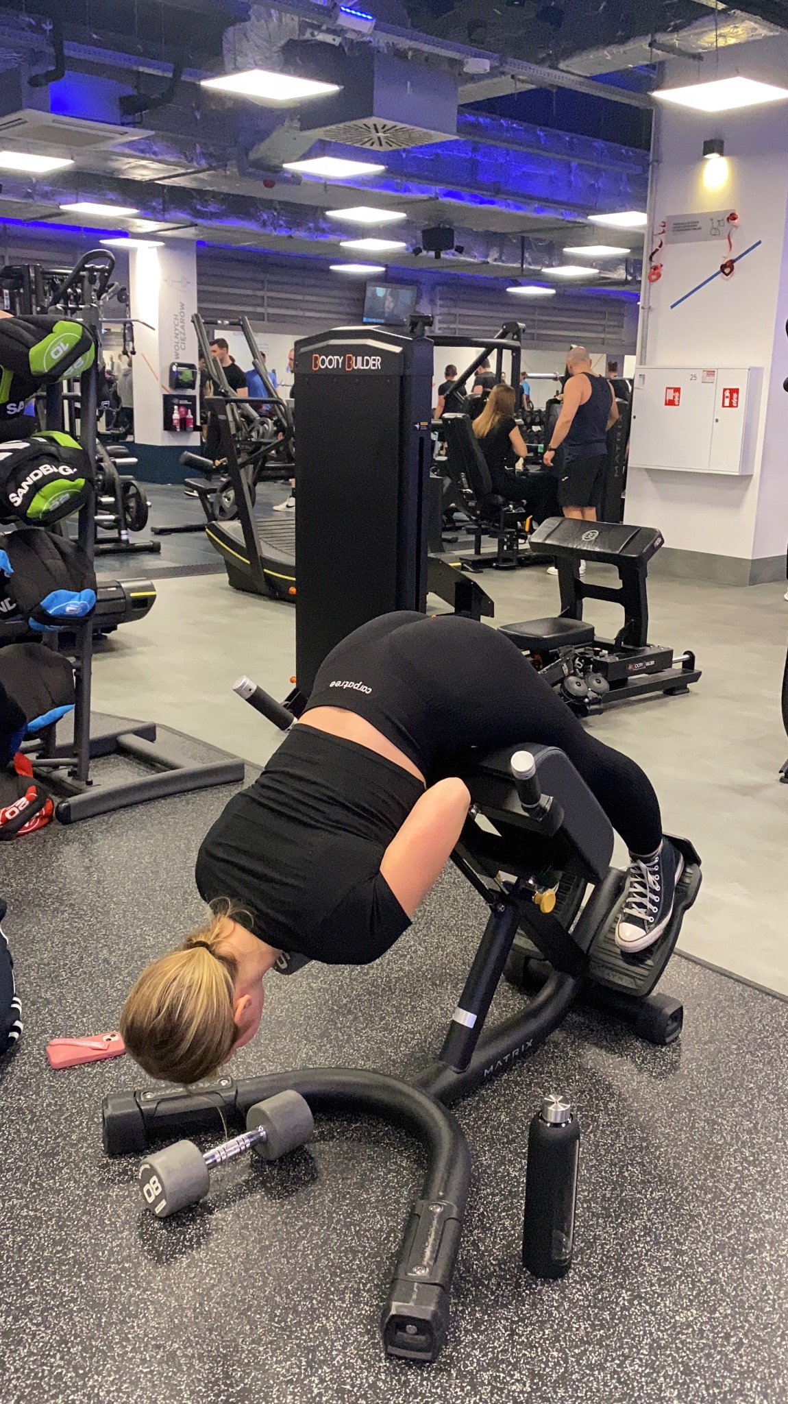 Kobieta w czarnym stroju sportowym wykonuje ćwiczenie na maszynie do wzmacniania mięśni grzbietu w siłowni, obok leżą hantle 8 kg i czarna butelka na wodę.