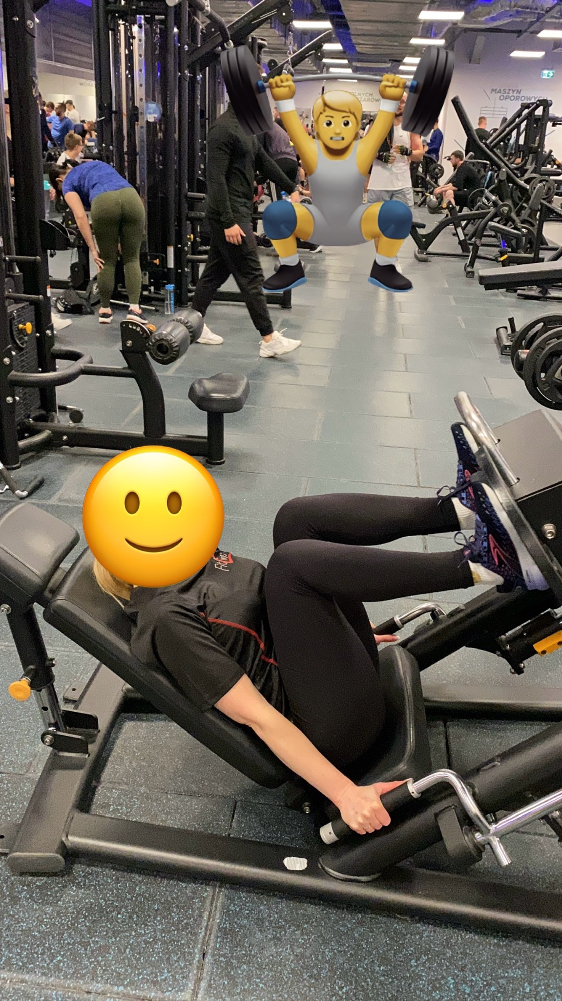 Kobieta ćwiczy na suwnicy na nogi w siłowni, w tle widoczni inni ćwiczący, podłoga pokryta niebieską gumą.